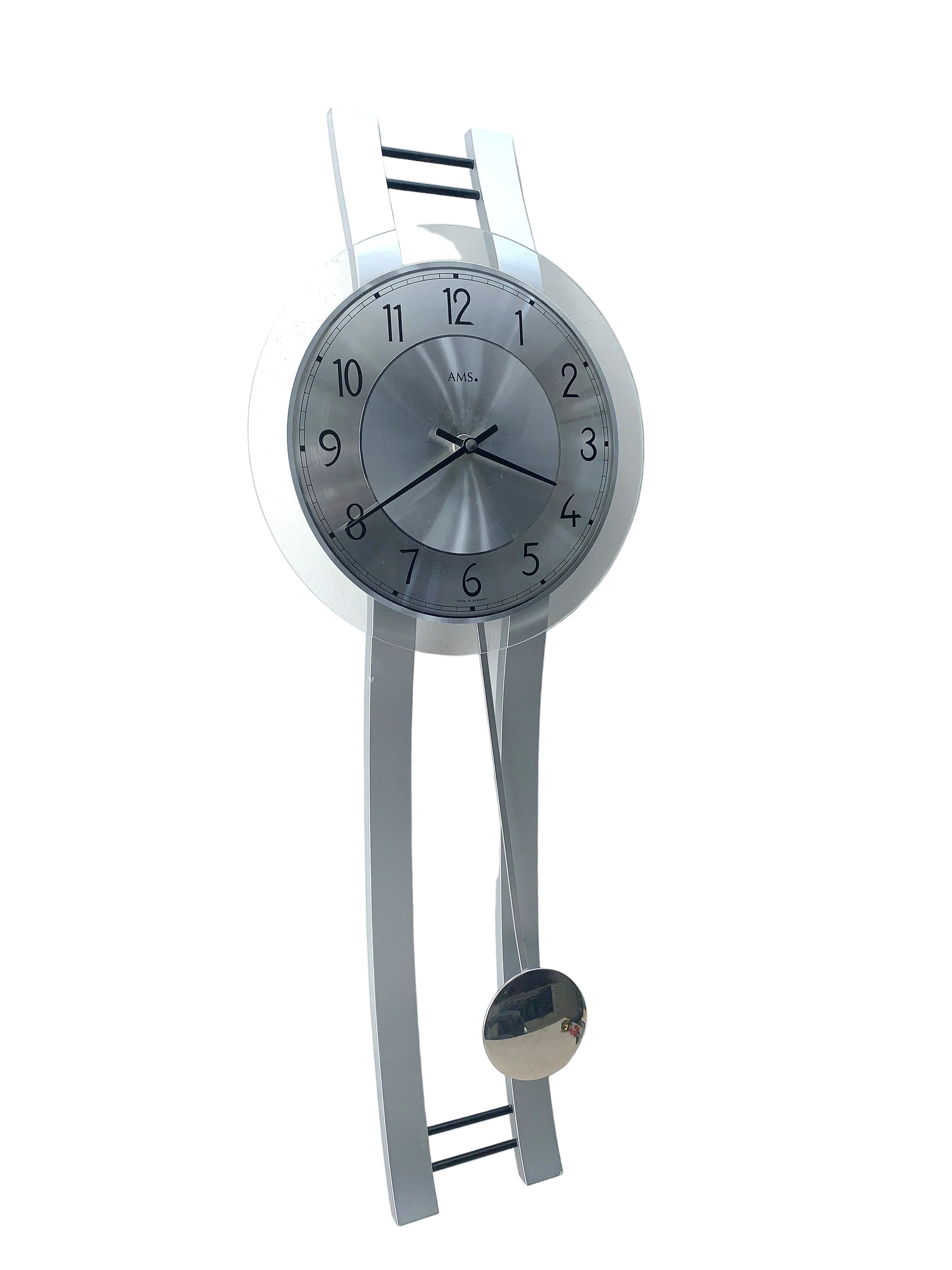 A modern wall clock with a silver metallic circular face and a matching silver pendulum, mounted on a white wall.
