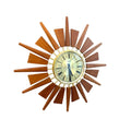 A mid-century modern sunburst wall clock with a warm brown wooden starburst frame and a cream-colored dial, mounted on a plain white wall.