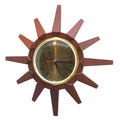 A mid-century modern sunburst wall clock with a dark reddish-brown geometric wooden frame, a gold-tone bezel, and a dark dial with black Roman numerals and a red second hand.