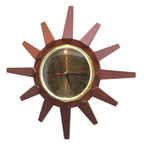 A mid-century modern sunburst wall clock with a dark reddish-brown geometric wooden frame, a gold-tone bezel, and a dark dial with black Roman numerals and a red second hand.