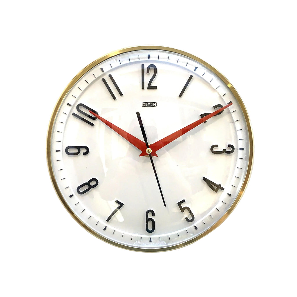 A round wall clock with a white face, black Arabic numerals, red hands, and a gold-toned metal frame, photographed against a plain off-white wall.
