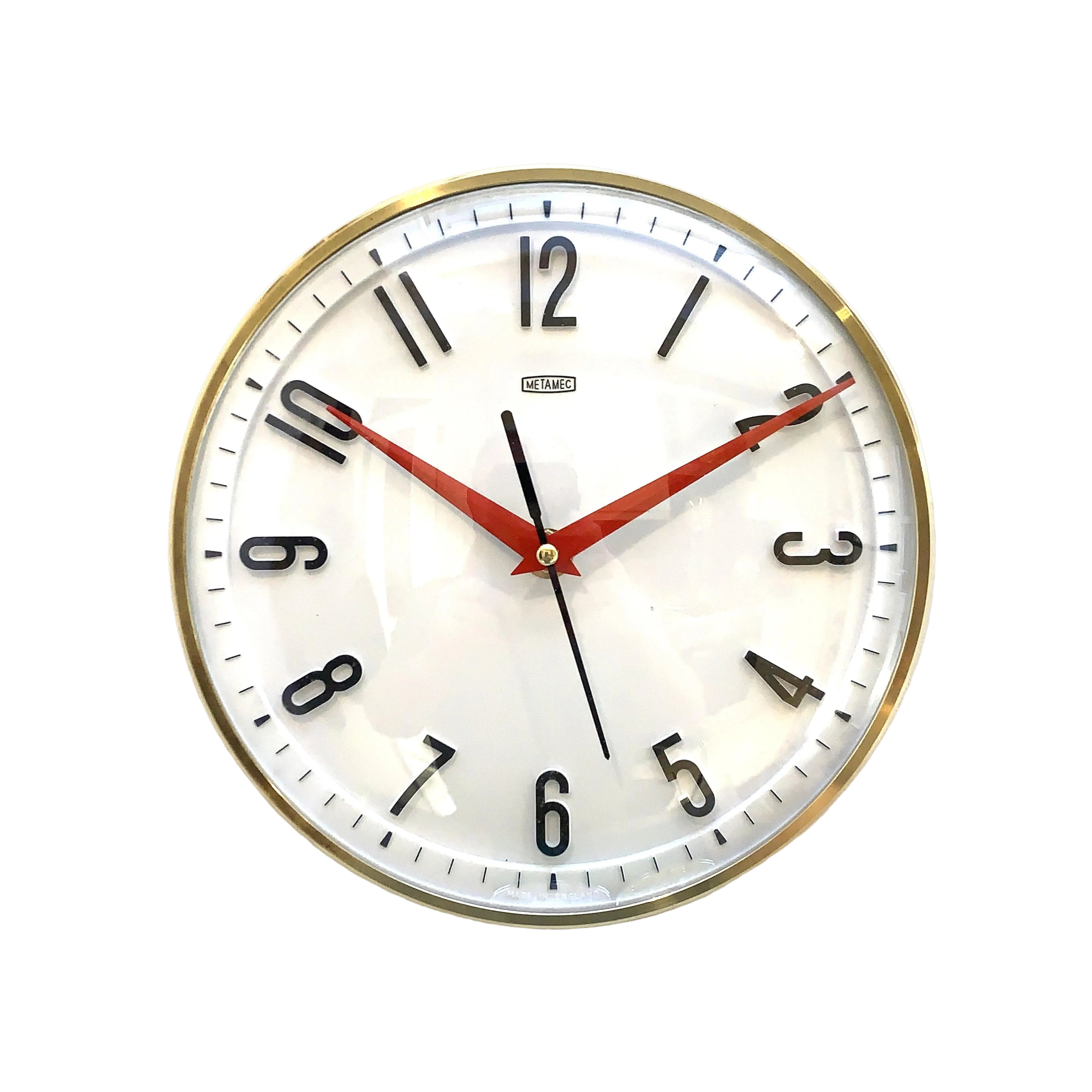 A round wall clock with a white face, black Arabic numerals, red hands, and a gold-toned metal frame, photographed against a plain off-white wall.