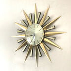 A mid-century modern sunburst wall clock with a gold-toned metal frame and radiating star-shaped hands, featuring a white clock face with black markings and a small calendar window at the top.