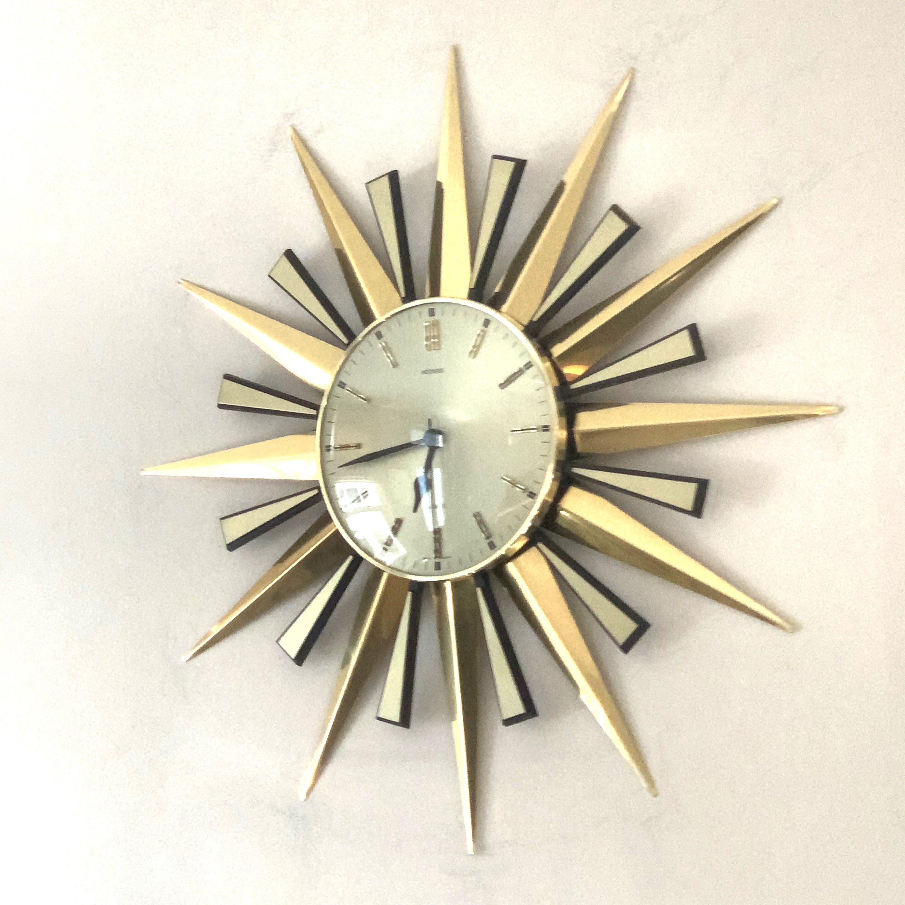 A mid-century modern sunburst wall clock with a gold-toned metal frame and radiating star-shaped hands, featuring a white clock face with black markings and a small calendar window at the top.