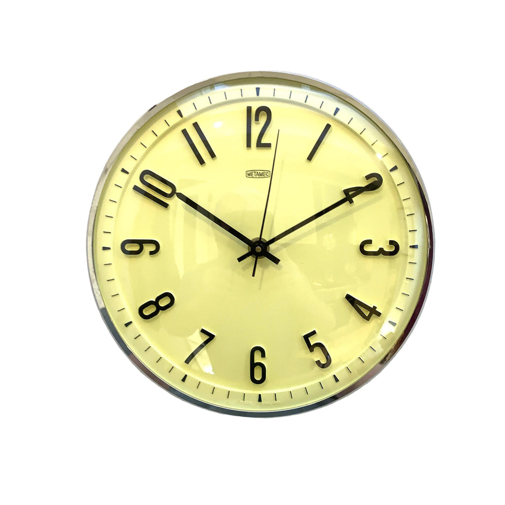 A round wall clock with a pale yellow dial, black Arabic numerals, black hands, and a silver-toned metal rim, mounted on a white wall.
