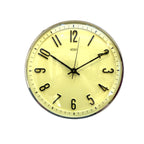 A round wall clock with a pale yellow dial, black Arabic numerals, black hands, and a silver-toned metal rim, mounted on a white wall.