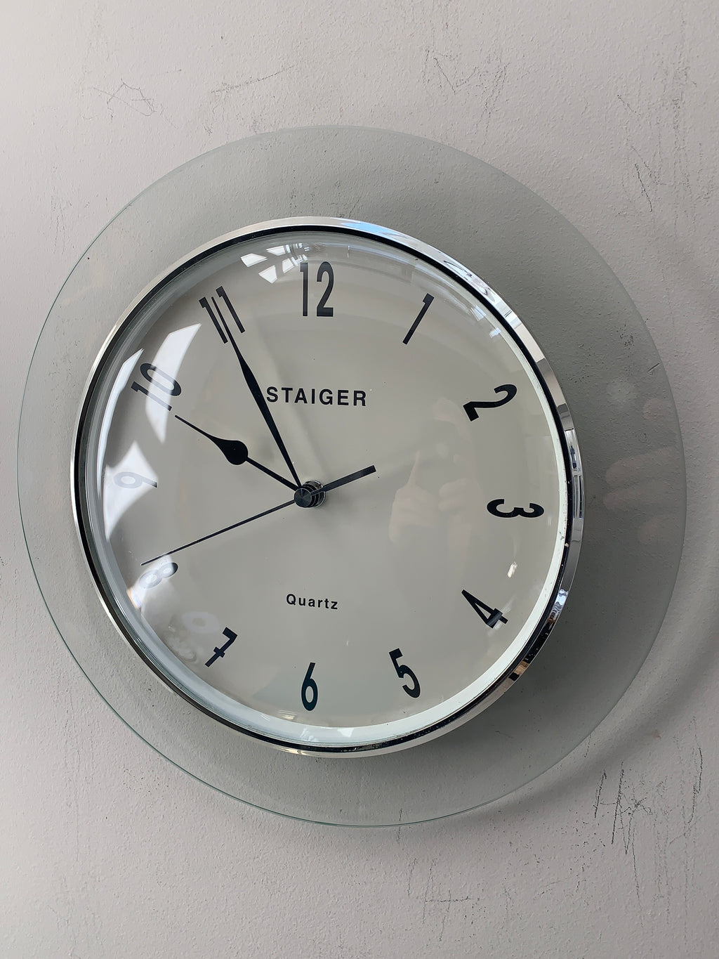 A round wall clock with a clear glass outer frame and a white dial, featuring black Arabic numerals and hands. The brand name 'STAIGER' is printed on the face, and the clock is mounted on a light gray textured wall.