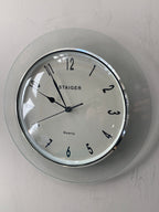 A round wall clock with a clear glass outer frame and a white dial, featuring black Arabic numerals and hands. The brand name 'STAIGER' is printed on the face, and the clock is mounted on a light gray textured wall.