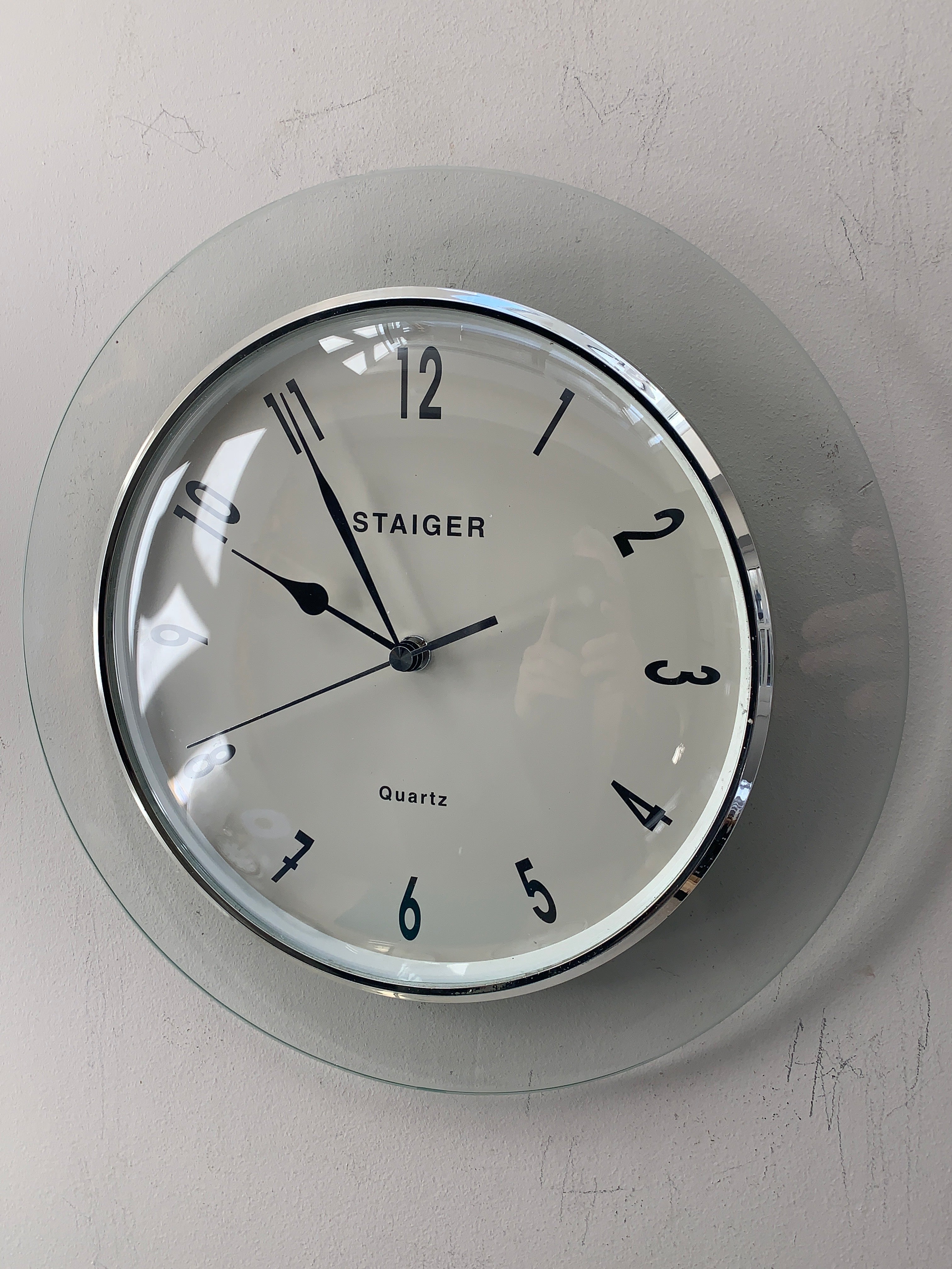 A round wall clock with a clear glass outer frame and a white dial, featuring black Arabic numerals and hands. The brand name 'STAIGER' is printed on the face, and the clock is mounted on a light gray textured wall.